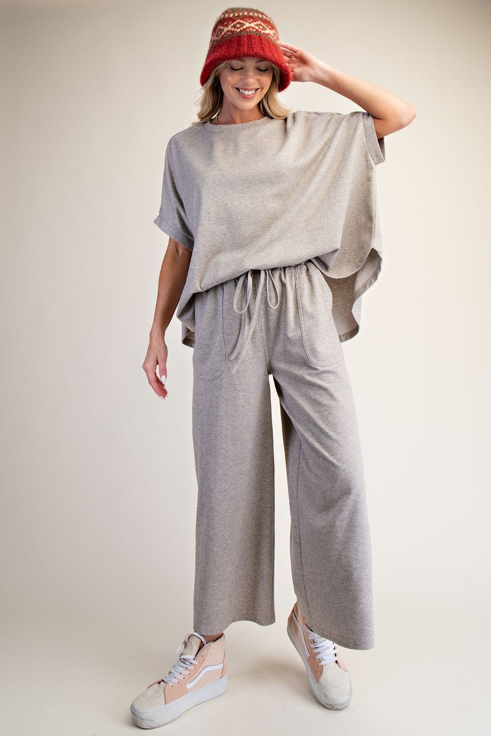 Woman wearing a gray outfit with wide-leg pants and a red hat on a plain background