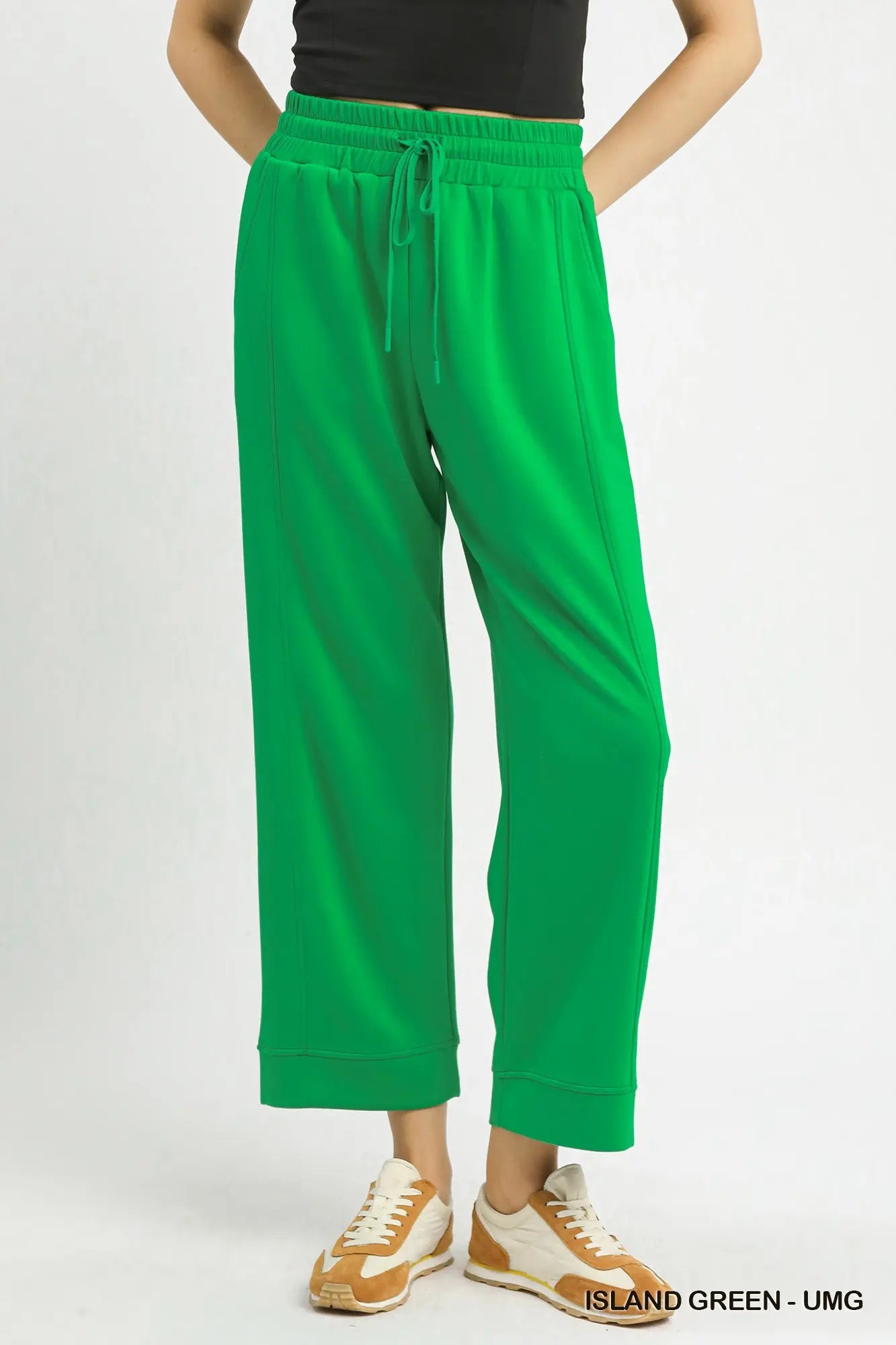 Bright green cropped sweatpants with an elastic drawstring waist, modeled with a black top and beige sneakers against a light gray studio background.