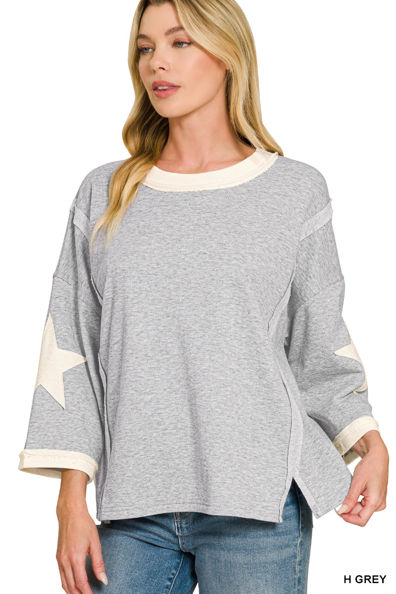 A woman wearing a heather gray long-sleeve top with cream-colored trim and a star patch on one sleeve, paired with blue jeans against a white background.