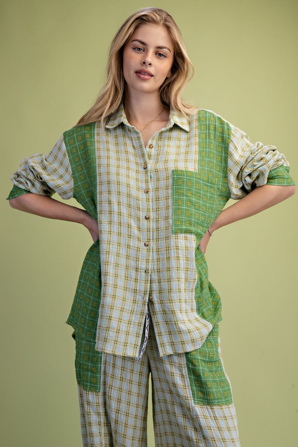 A woman modeling a patchwork button-up shirt and matching pants in green and beige plaid fabrics, posed against a solid light green studio background.