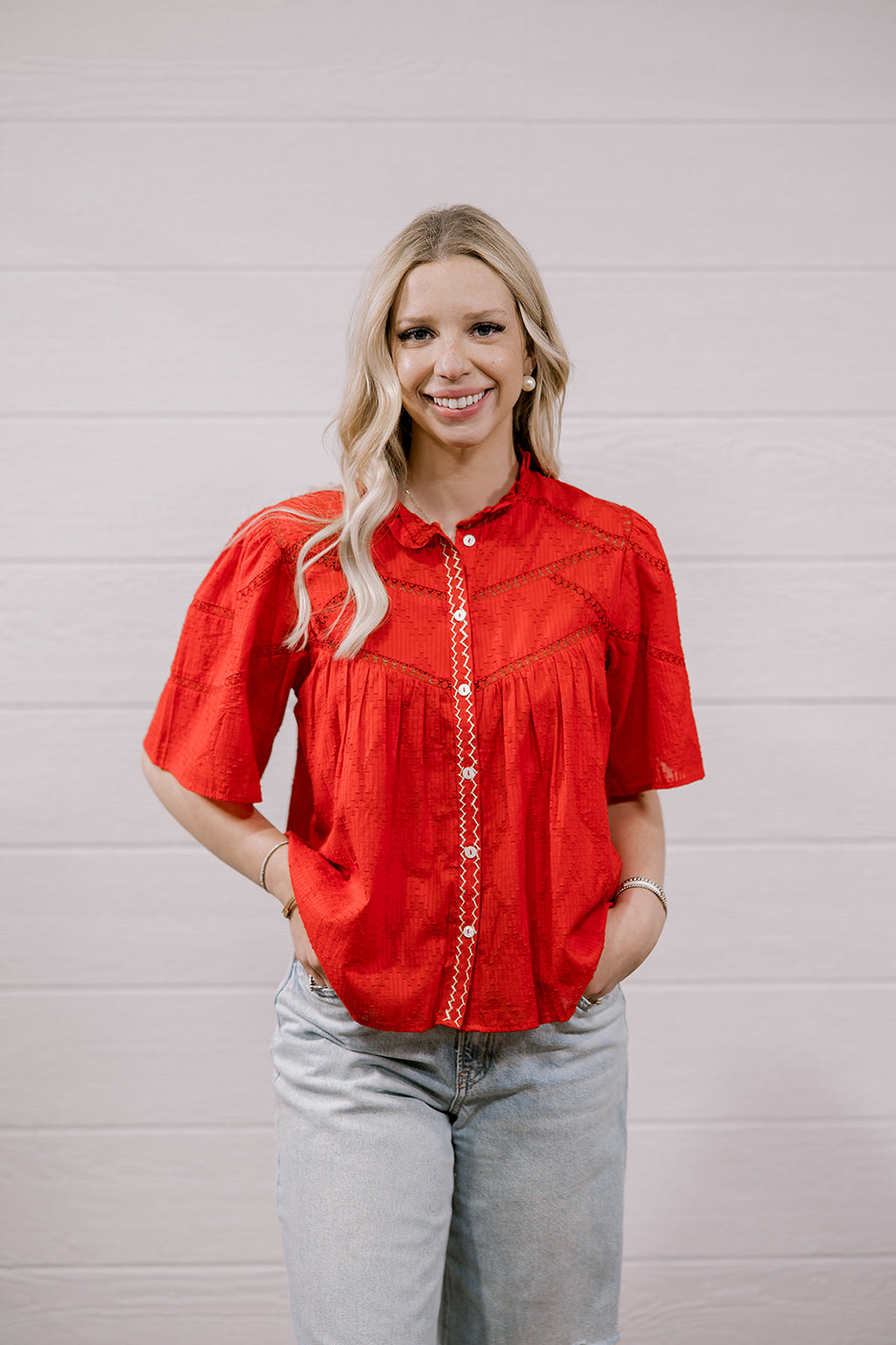 A woman with blonde hair wearing a bright red short-sleeve embroidered blouse and light-wash jeans, standing against a white paneled wall.