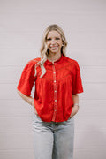 A woman with blonde hair wearing a bright red short-sleeve embroidered blouse and light-wash jeans, standing against a white paneled wall.