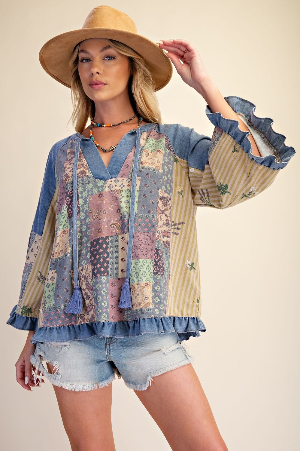 A woman modeling a bohemian-style patchwork blouse with blue denim sleeves and ruffles, paired with denim shorts and a wide-brimmed straw hat against a solid beige background.
