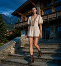 Woman in a beige dress with brown embroidery standing on stone steps in front of a wooden building with mountains in the background.