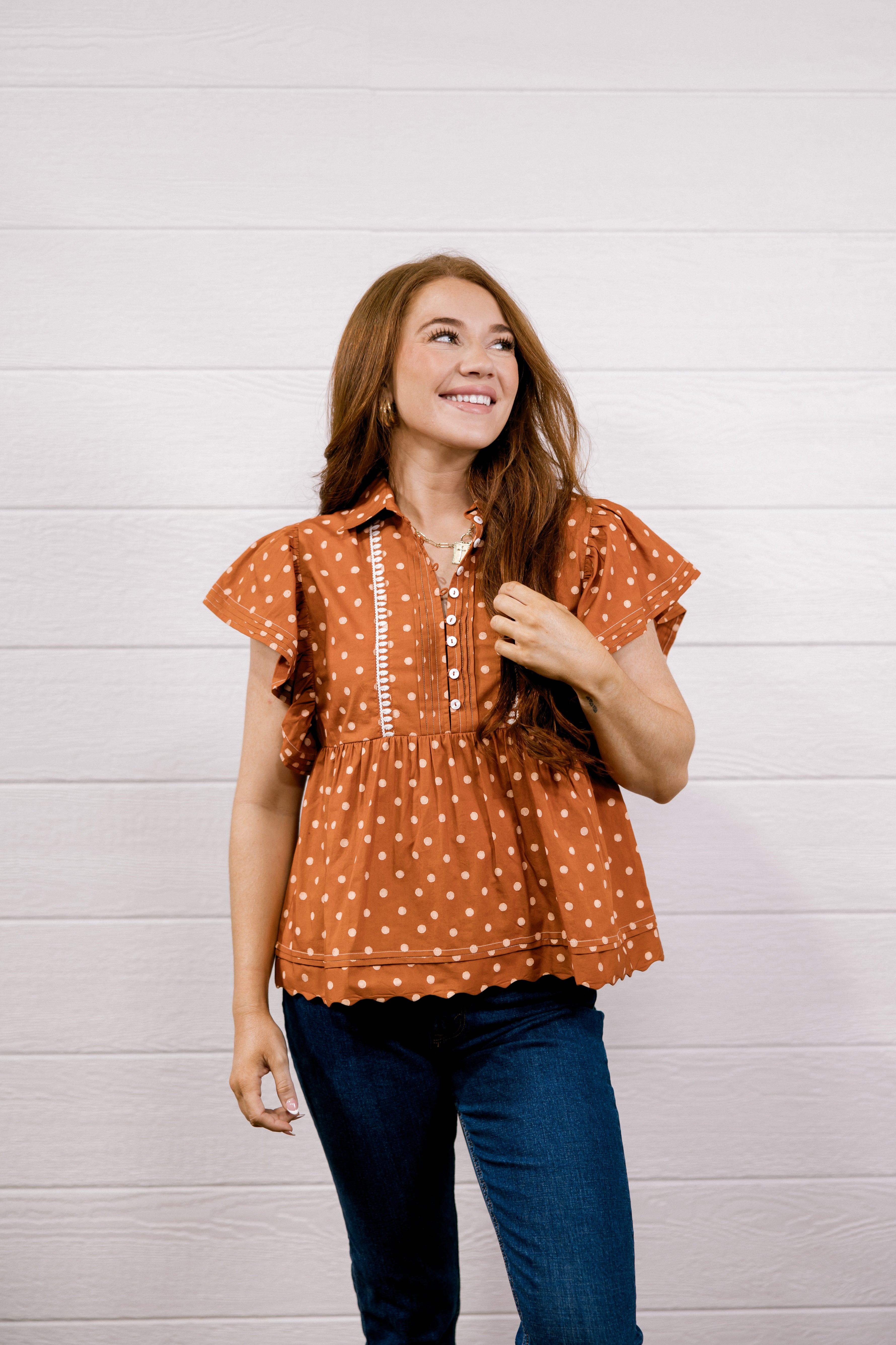 A woman wearing a rust-orange short-sleeve blouse with white polka dots and a white vertical zipper detail on the chest, paired with dark blue jeans, standing against a white wood-paneled wall.