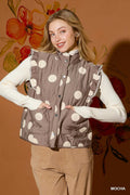 A woman models a sleeveless quilted vest with a brown and cream polka dot pattern, layered over a white long-sleeve top and tan pants, set against a warm orange floral wall.