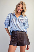 A woman modeling a short-sleeve button-up shirt with a blue and white polka dot pattern, paired with dark brown shorts, against a light gray background.