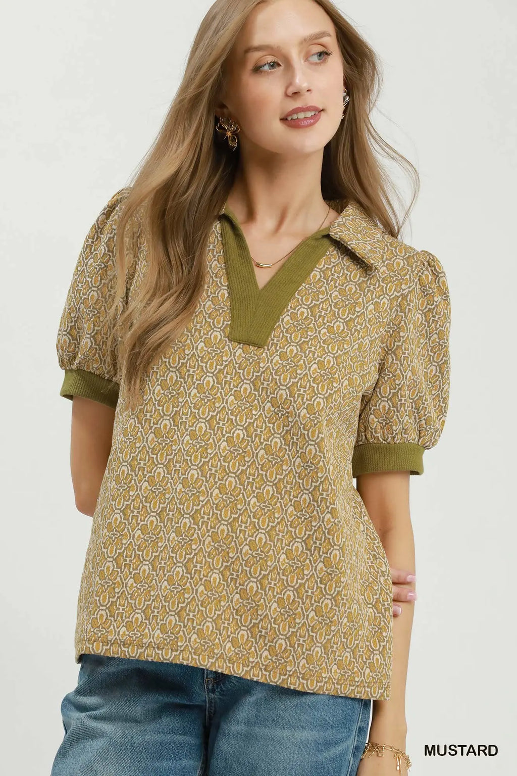 A woman wearing a mustard yellow patterned short-sleeve top with olive green trim and a V-neckline, paired with blue jeans, against a plain light background.