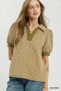 A woman wearing a mustard yellow patterned short-sleeve top with olive green trim and a V-neckline, paired with blue jeans, against a plain light background.
