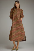 A woman modeling a brown midi dress with a red and green scalloped pattern, styled with olive green pointed flats against a neutral gray studio background.