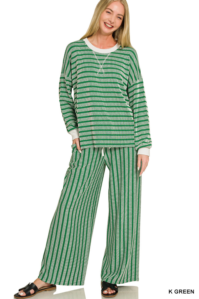 A woman modeling a two-piece lounge set consisting of a long-sleeve top and matching wide-leg pants, both in a green and white striped pattern. She is wearing black slide sandals against a plain white background.