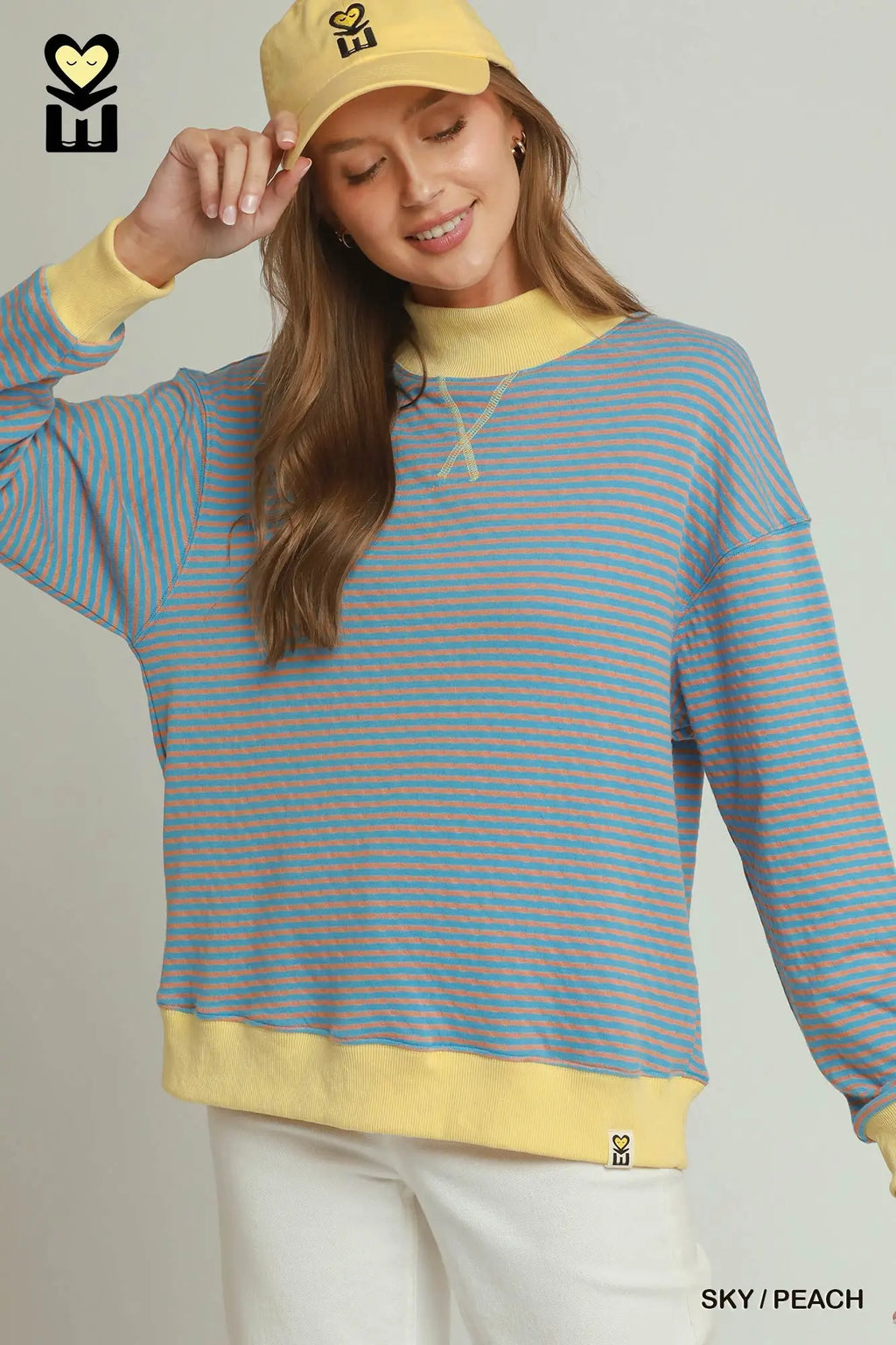 A woman wearing a light blue and peach striped crewneck sweatshirt with yellow ribbed trim, paired with a matching yellow baseball cap and white pants, against a light gray studio background.