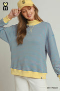 A woman wearing a light blue and peach striped crewneck sweatshirt with yellow ribbed trim, paired with a matching yellow baseball cap and white pants, against a light gray studio background.