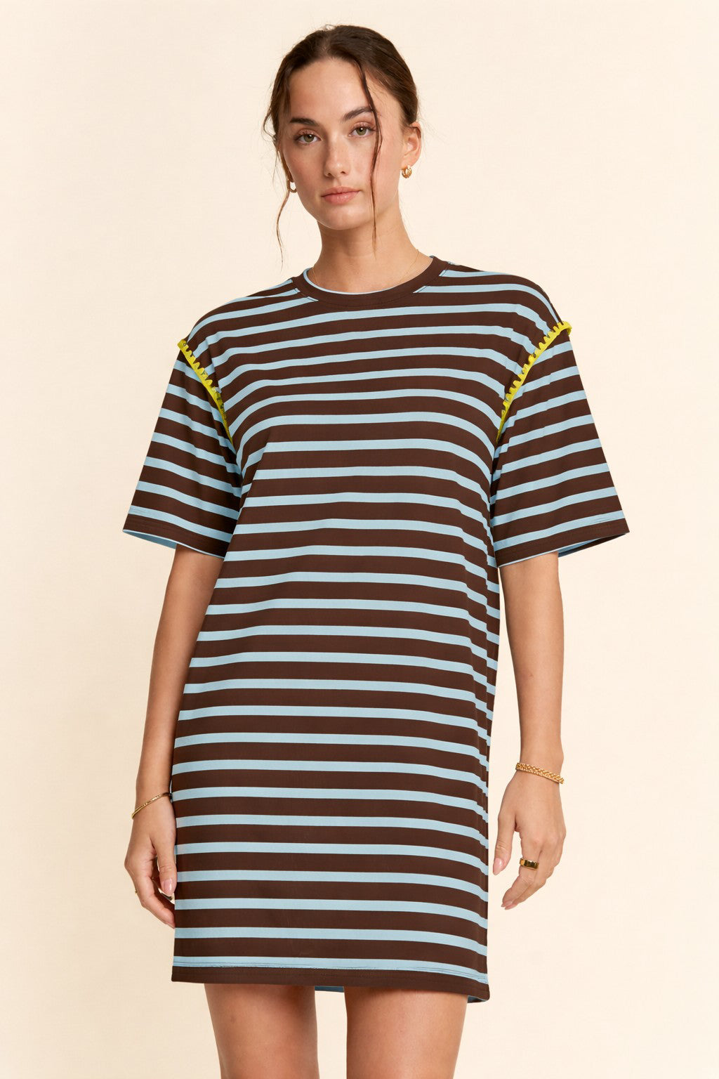 A woman modeling an oversized short-sleeve t-shirt dress with horizontal brown, black, and light blue stripes, featuring yellow embroidered details on the shoulders, set against a solid beige background.