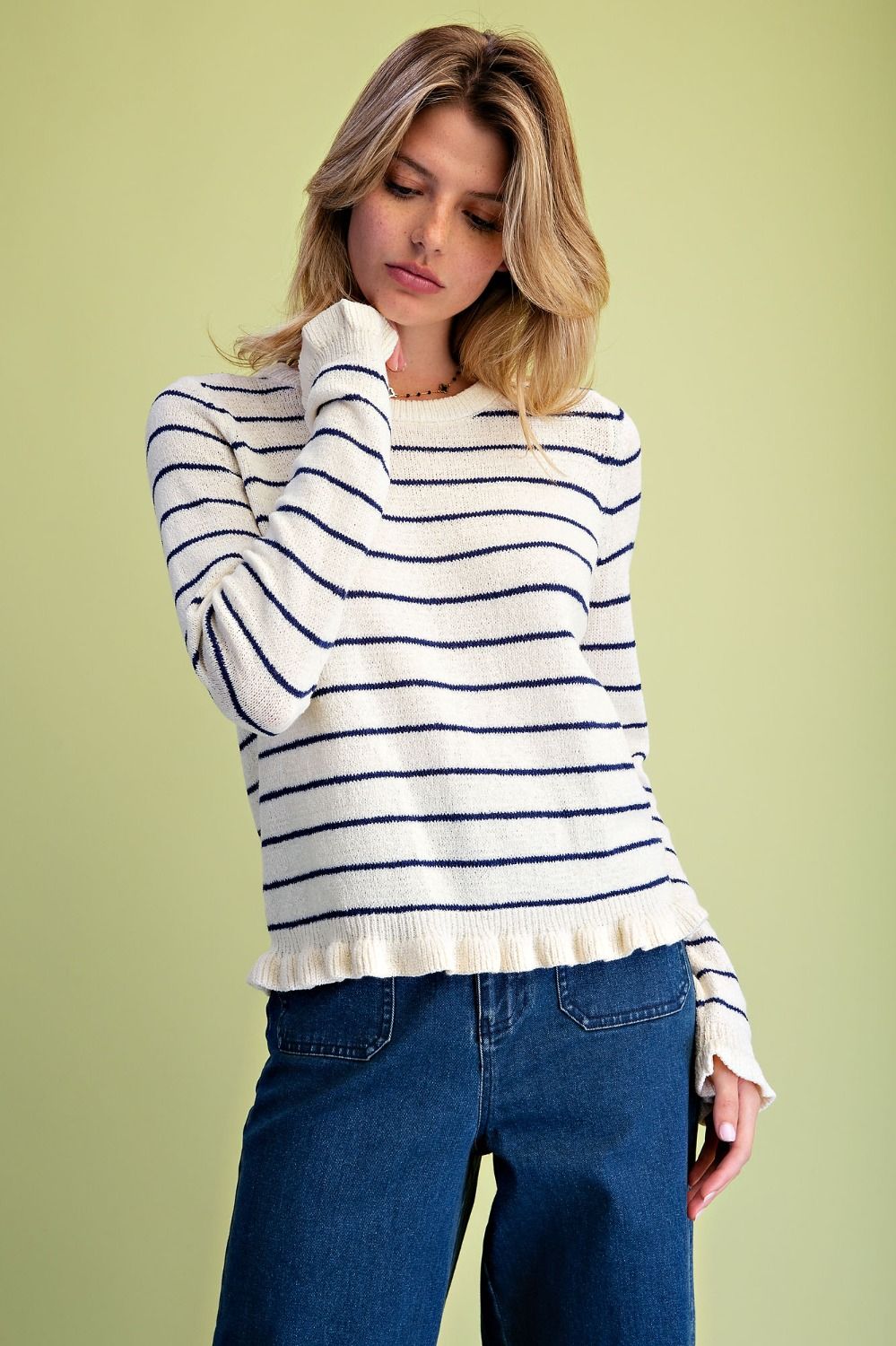 A woman wearing a cream and navy blue horizontally striped knit sweater with a ruffled hem, paired with blue jeans, set against a solid light olive green background.