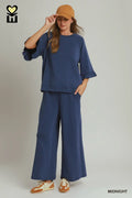 A woman modeling a navy blue two-piece lounge set consisting of a short-sleeve top with flared sleeves and matching wide-leg pants, styled with a tan cap, brown and cream sneakers, and a brand logo in the top left corner.