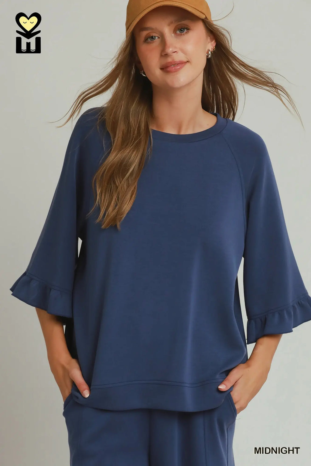 A woman wearing a navy blue lounge set consisting of a short-sleeve top with ruffled cuffs and matching pants, paired with a tan baseball cap. The text 'MIDNIGHT' is visible in the bottom right corner.