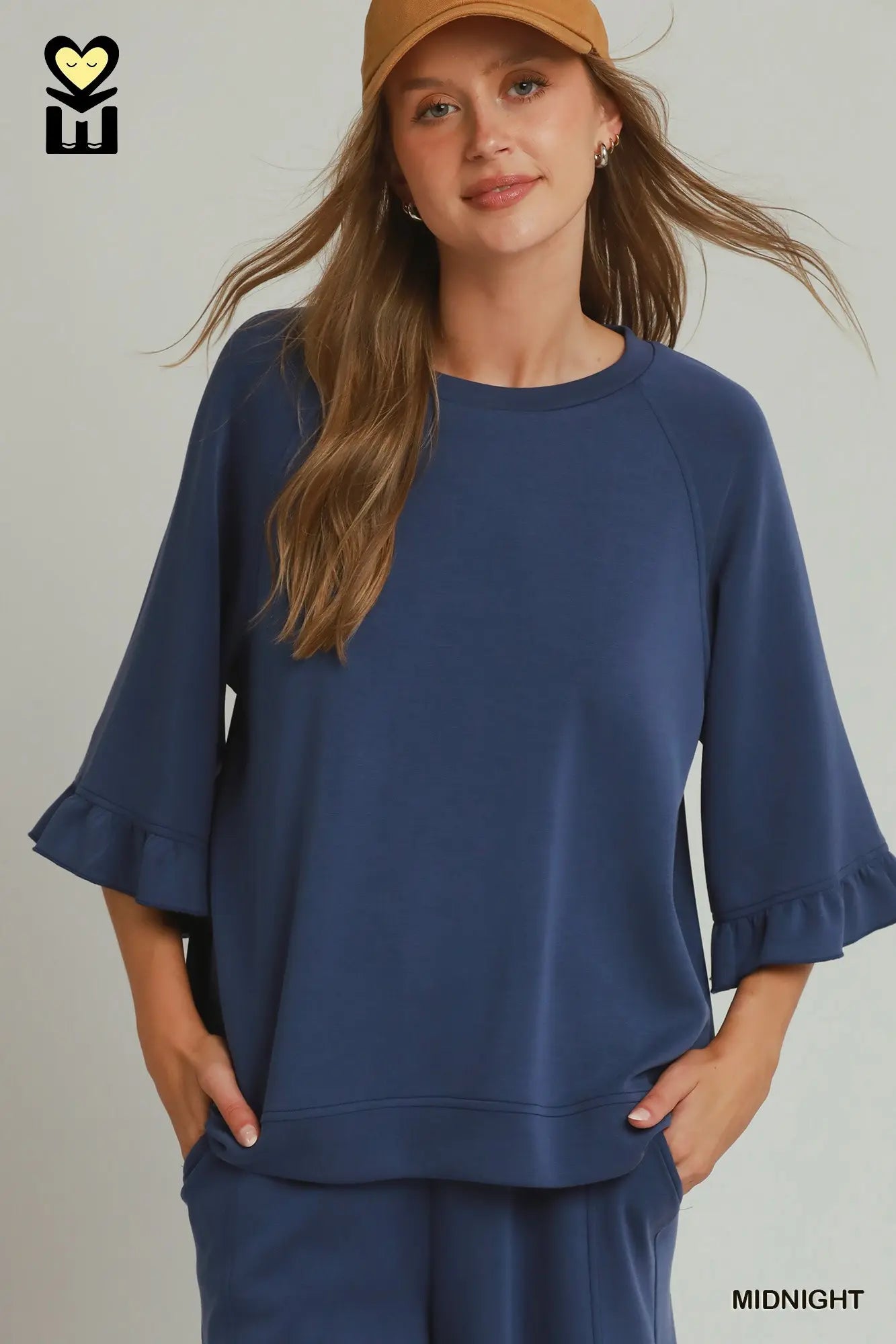 A woman wearing a navy blue lounge set consisting of a short-sleeve top with ruffled cuffs and matching pants, paired with a tan baseball cap. The text 'MIDNIGHT' is visible in the bottom right corner.