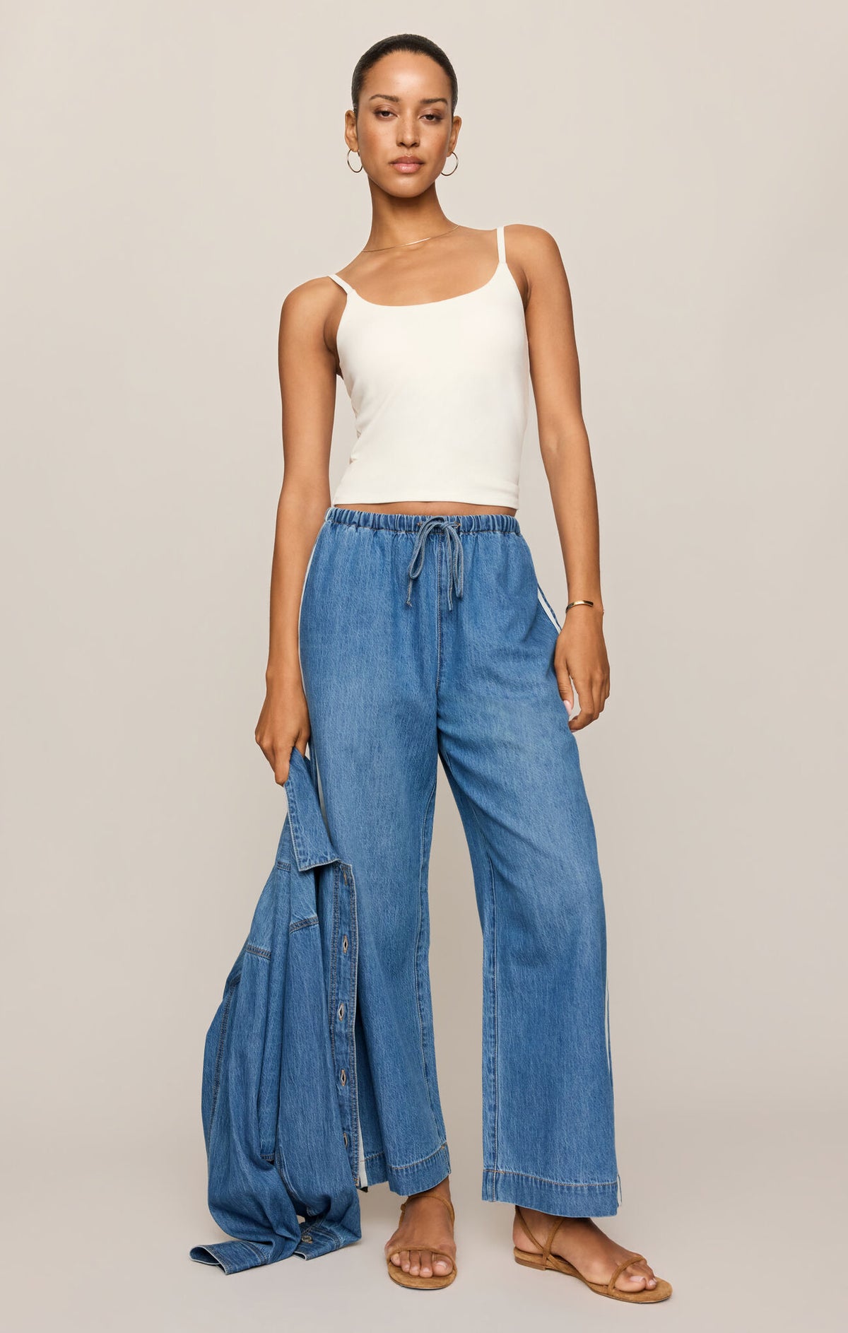Woman wearing a white tank top and blue wide-leg jeans on a beige background