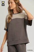 A woman wearing a color-block short-sleeve top in charcoal and beige, paired with matching charcoal pants, against a light gray studio background.