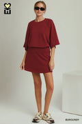 A woman modeling a burgundy two-piece lounge set consisting of a short-sleeve top and a matching mini skirt, styled with beige sneakers and sunglasses against a light gray studio background.