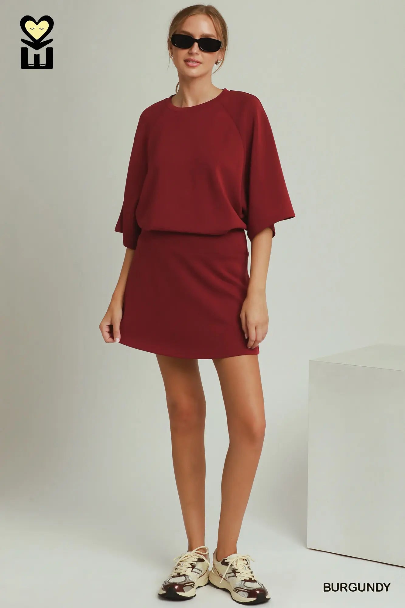 A woman modeling a burgundy two-piece lounge set consisting of a short-sleeve top and a matching mini skirt, styled with beige sneakers and sunglasses against a light gray studio background.