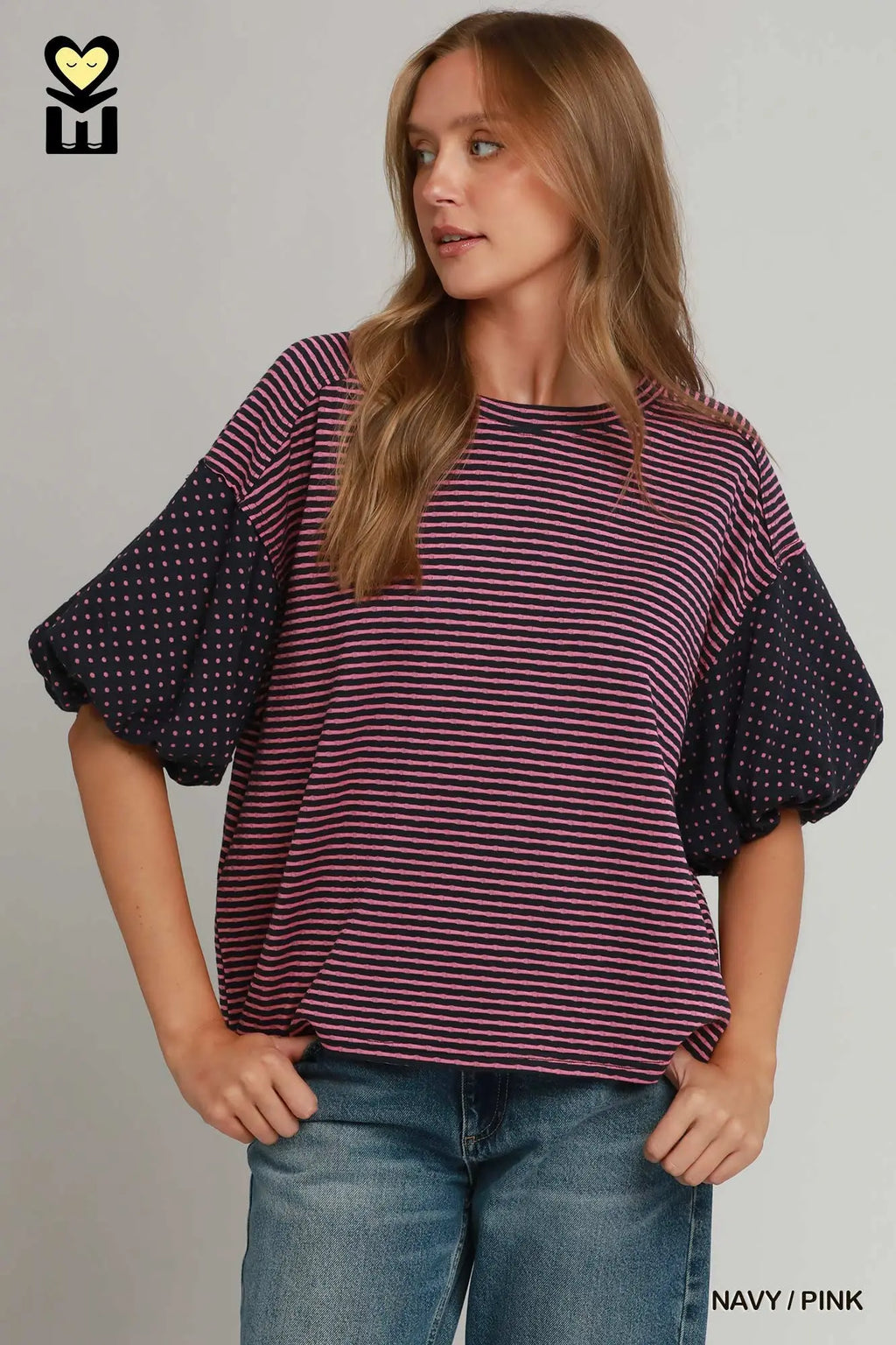 A woman wearing a short-sleeve top with a pink and dark red striped body and navy polka-dot balloon sleeves, paired with blue jeans, against a light gray background.