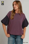 A woman wearing a short-sleeve top with a pink and dark red striped body and navy polka-dot balloon sleeves, paired with blue jeans, against a light gray background.