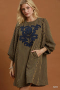 A woman models a loose-fitting, knee-length dress with vertical navy and olive-brown stripes, featuring a large navy floral embroidery on the chest and elbow-length puff sleeves. She is posed against a solid warm brown studio backdrop.