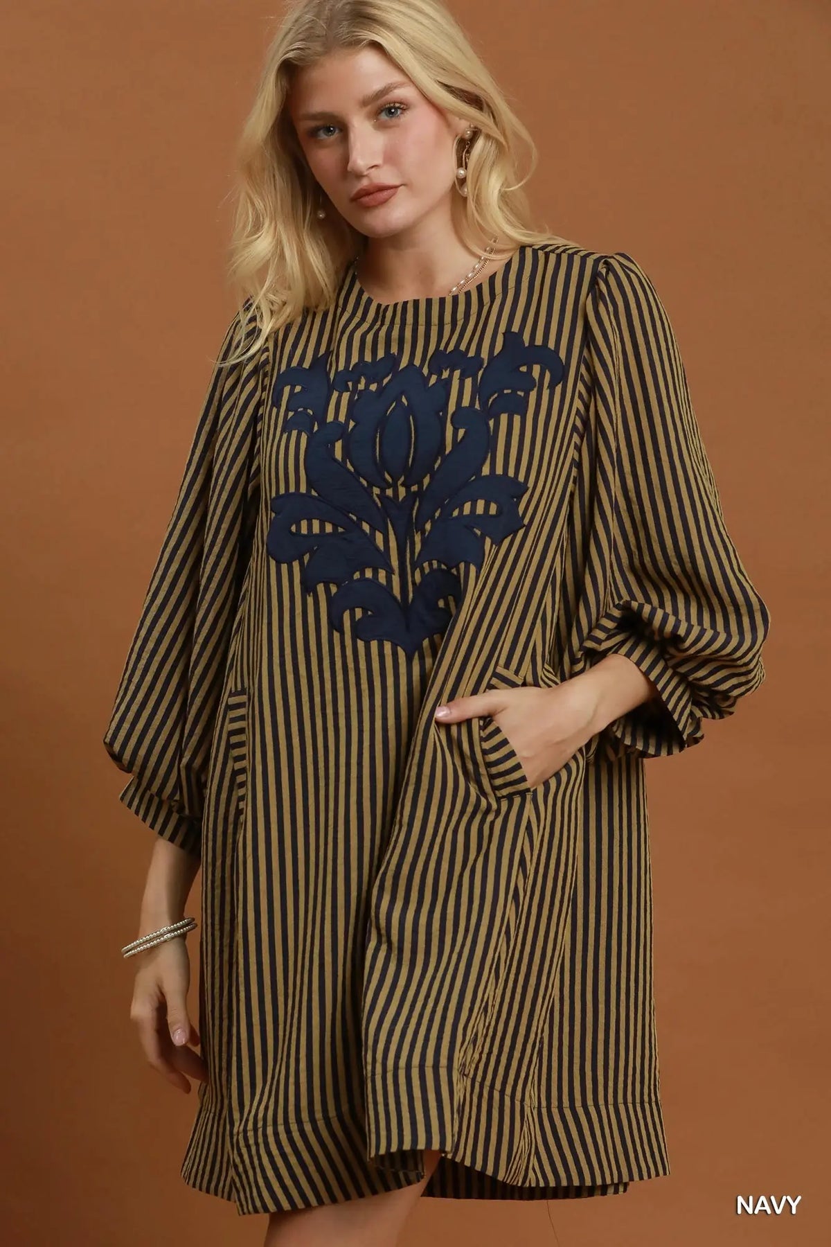 A woman models a loose-fitting, knee-length dress with vertical navy and olive-brown stripes, featuring a large navy floral embroidery on the chest and elbow-length puff sleeves. She is posed against a solid warm brown studio backdrop.