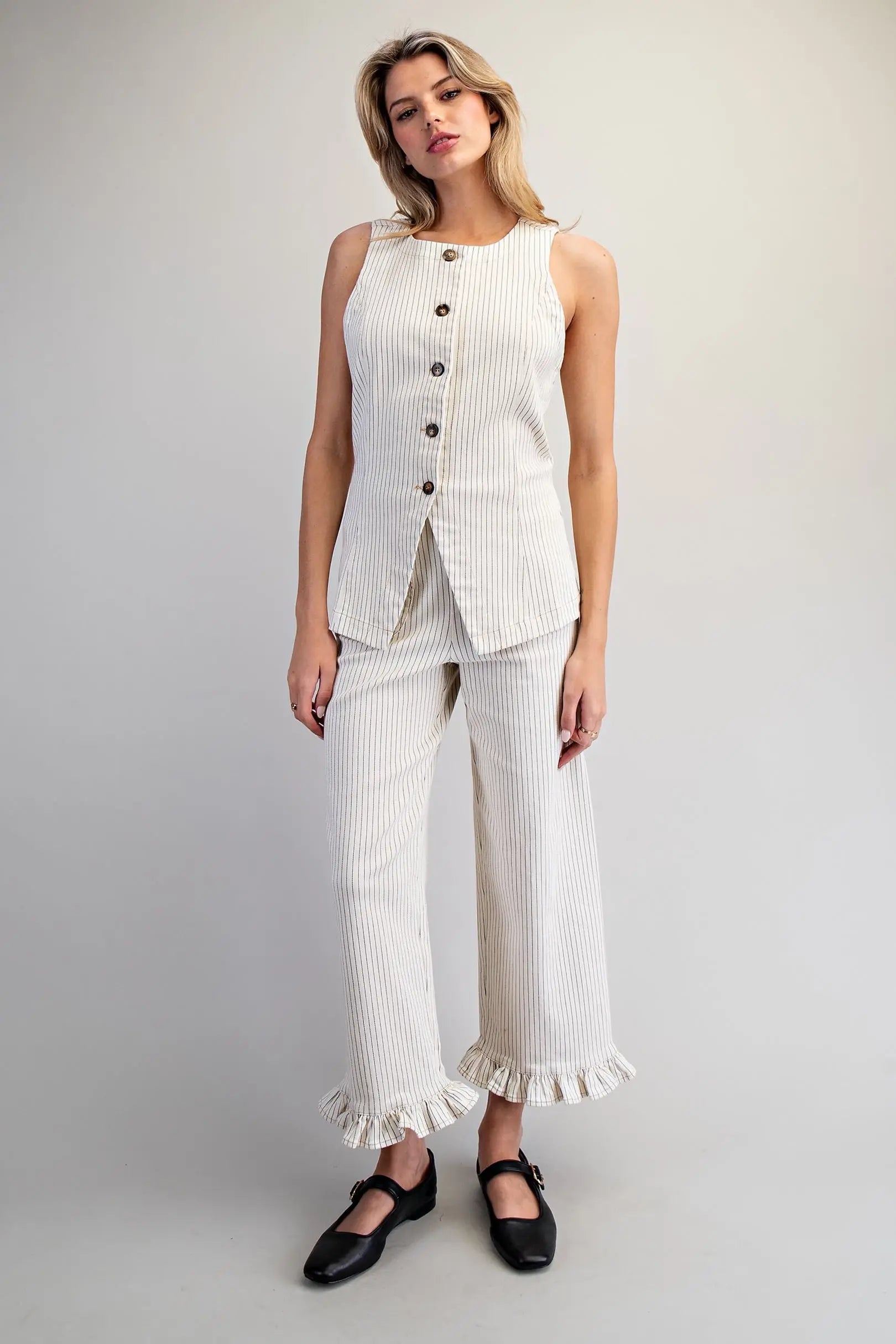 Woman wearing a white sleeveless top and pants with buttons on a plain background