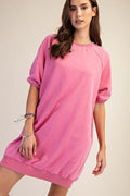 A woman wearing a pink short-sleeve crewneck sweatshirt dress with a relaxed, oversized fit and ribbed hem, photographed against a plain beige studio background.