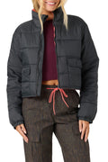 A woman wearing a cropped black puffer jacket with a high collar, zippered front, and flap pockets, styled over a burgundy crop top and brown patterned pants with a coral drawstring, against a plain white background.
