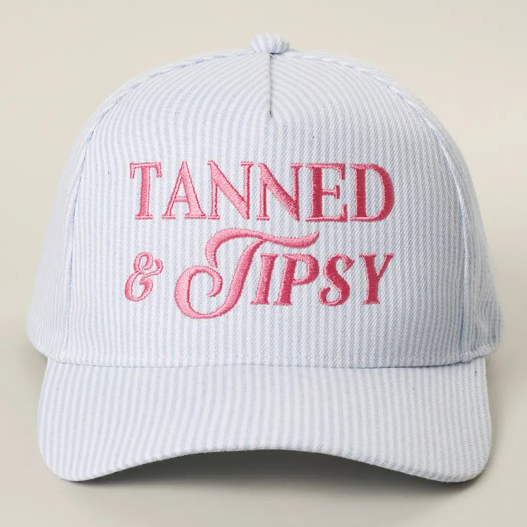 Light blue cap with pink 'Tanned & Tipsy' text on a beige background