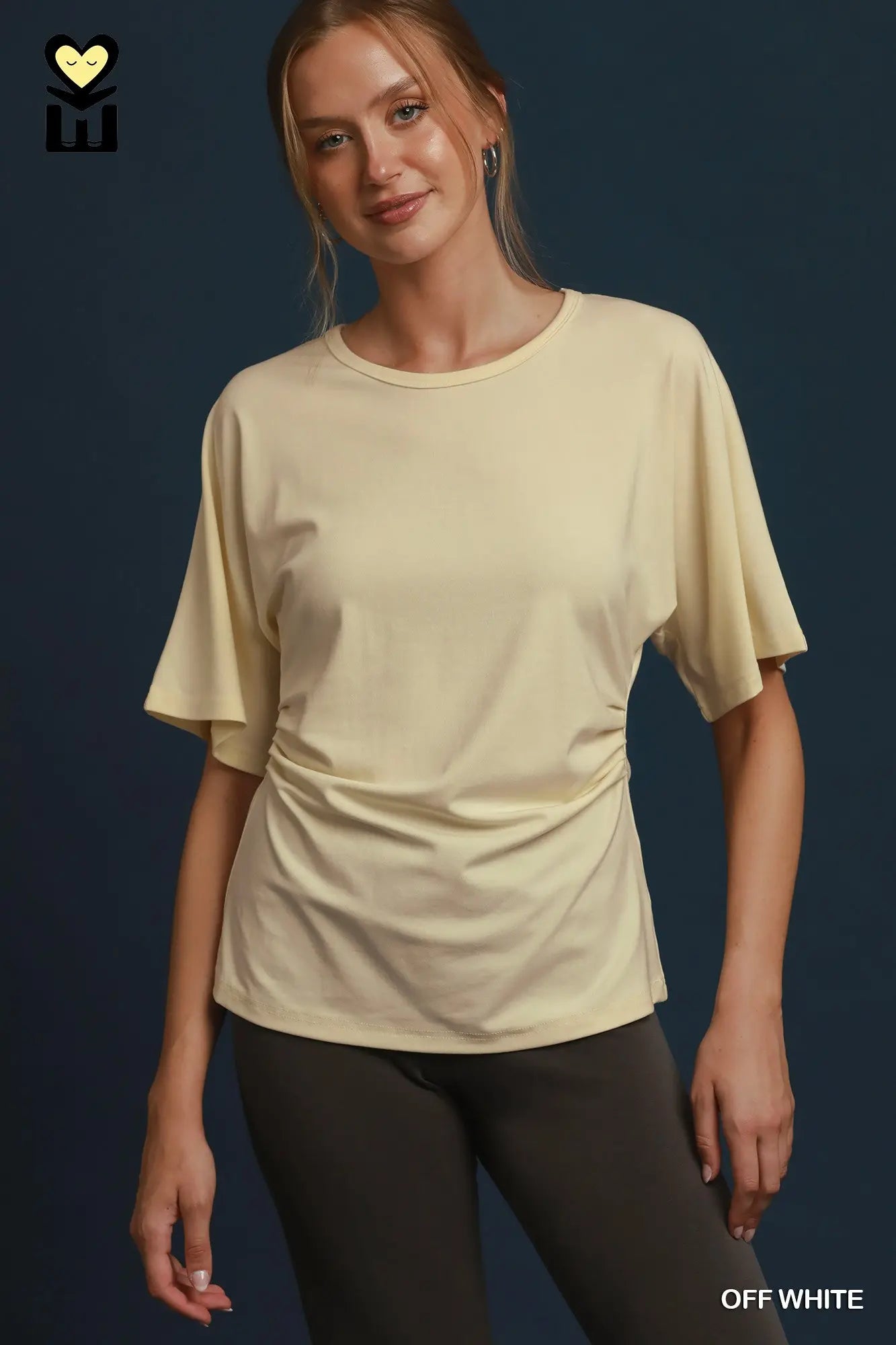 A woman modeling an off-white short-sleeve crewneck t-shirt with a gathered side detail, paired with dark gray leggings, against a dark blue background.