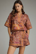 A woman modeling a matching two-piece outfit with a brown base and a vibrant orange, pink, and yellow floral pattern. The set includes a short-sleeve button-up shirt and a matching mini skirt, photographed against a solid gray studio background.