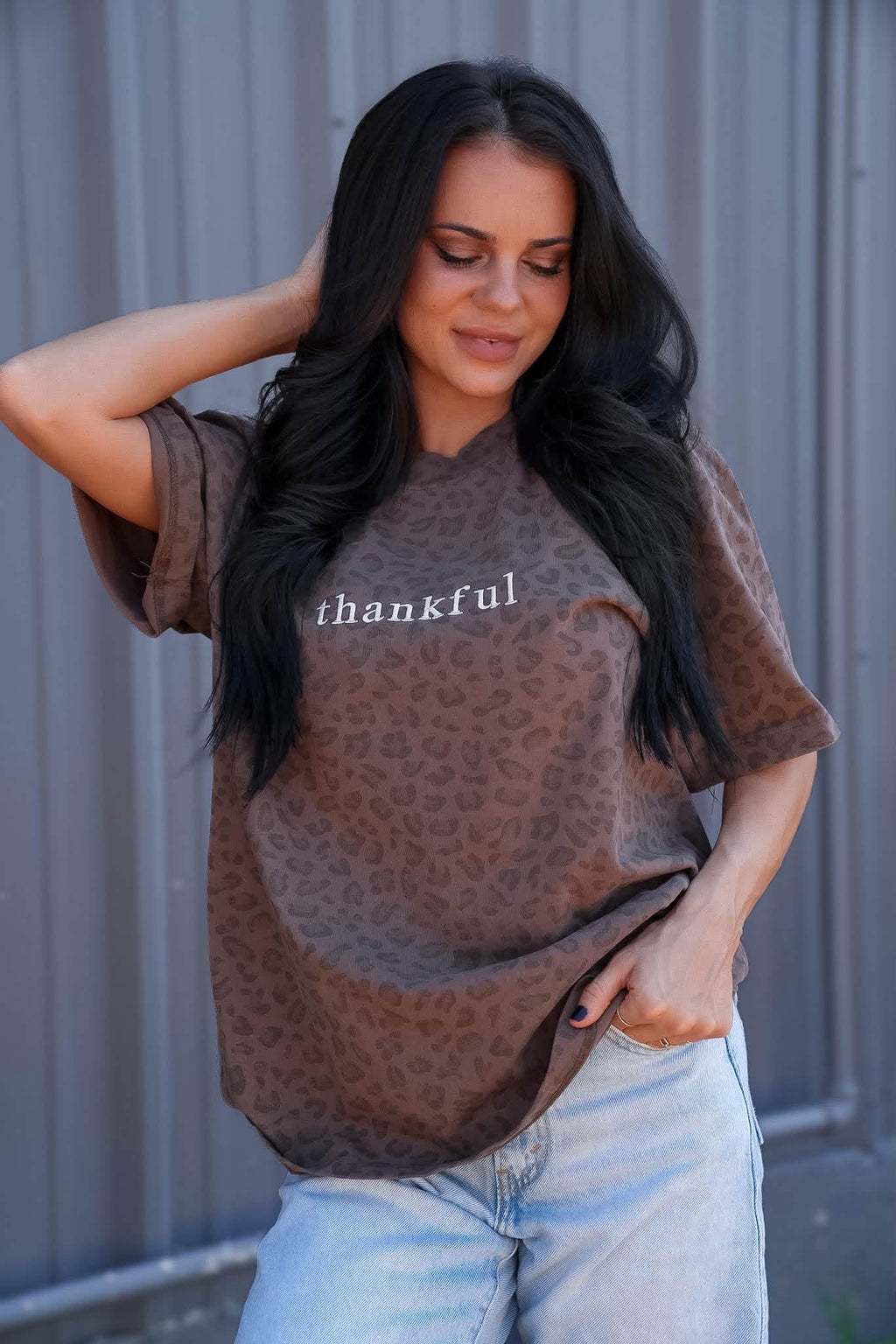 A woman wearing a brown leopard-print t-shirt with the word 'thankful' printed in white across the chest, paired with light-wash jeans, posing in front of a gray corrugated metal wall.