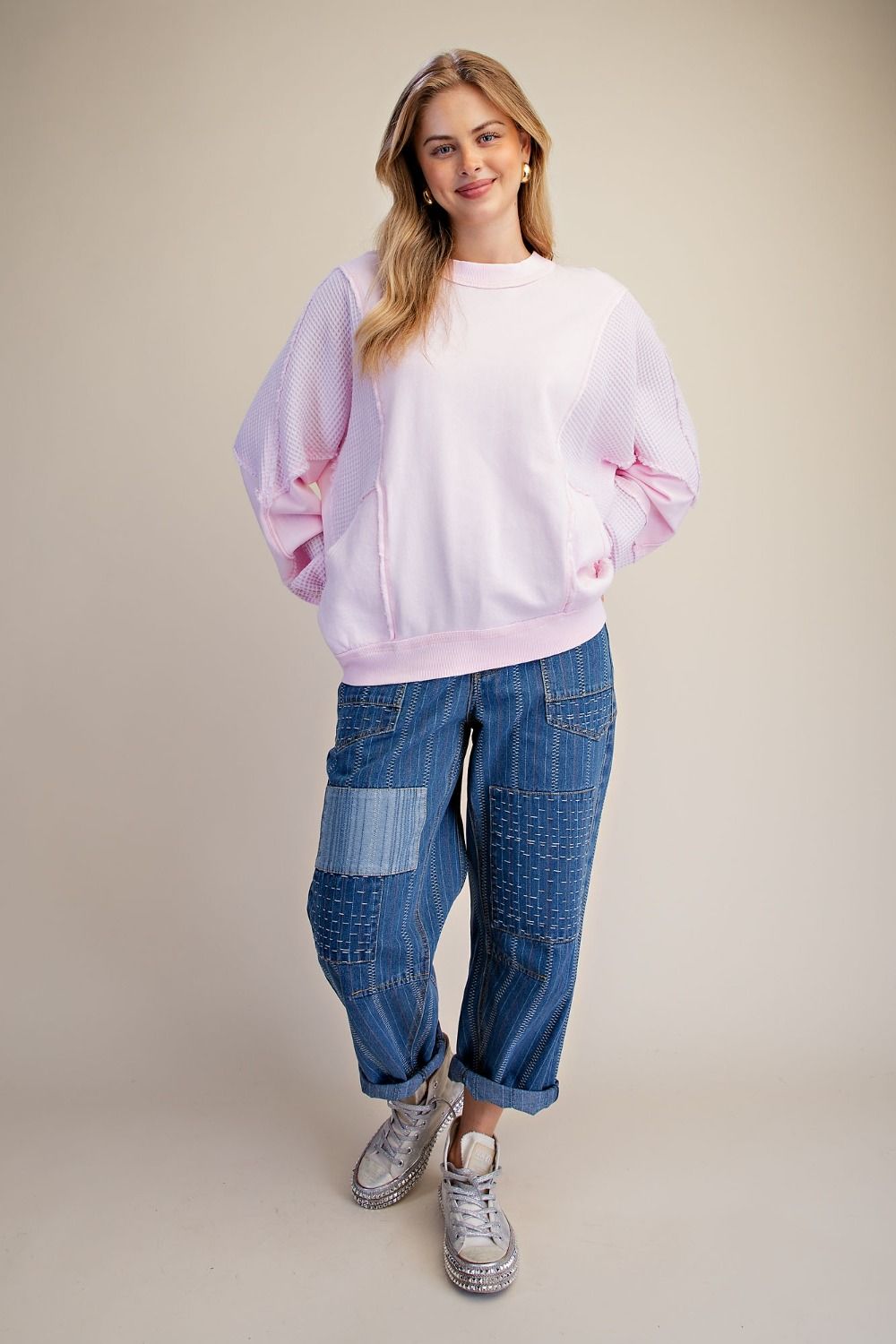 A woman modeling a light pink crewneck sweatshirt and blue patchwork denim jeans with gray sneakers, standing against a solid beige studio background.