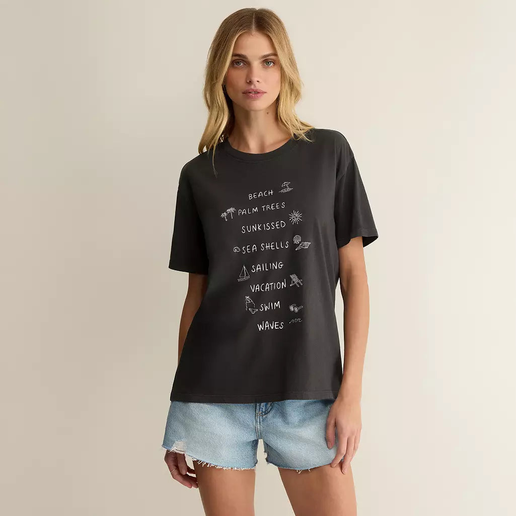 A woman modeling a black graphic t-shirt with white hand-drawn style text and illustrations, paired with light-wash denim shorts, against a solid light beige background.