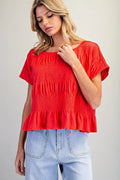 A woman wearing a bright red short-sleeve smocked blouse with a ruffled hem, paired with light blue jeans, photographed against a plain light gray studio background.