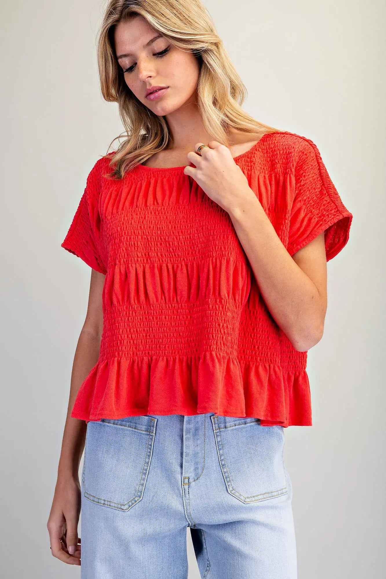 A woman wearing a bright red short-sleeve smocked blouse with a ruffled hem, paired with light blue jeans, photographed against a plain light gray studio background.