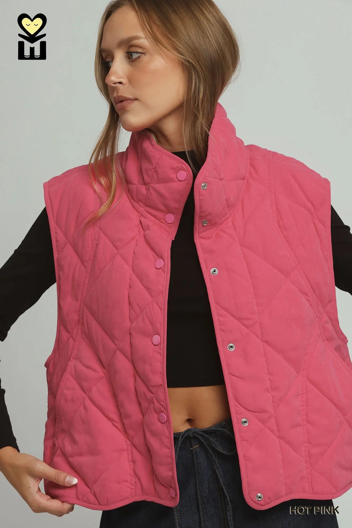A woman wearing a quilted sleeveless puffer vest in hot pink over a black cropped top and dark jeans, shown against a plain light gray studio background.