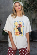 A woman wearing an oversized cream-colored graphic t-shirt with a dachshund print, paired with red and white polka dot pants and a white lace headband, standing in an outdoor patio setting.