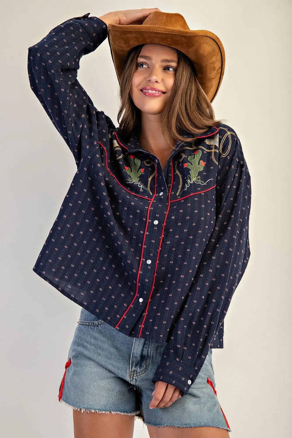 Woman wearing a navy embroidered shirt and denim shorts, holding a brown cowboy hat.