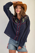 Woman wearing a navy embroidered shirt and denim shorts, holding a brown cowboy hat.