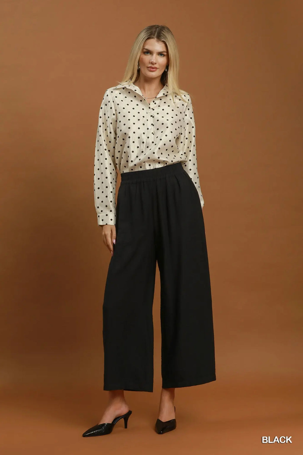 A woman modeling a cream long-sleeve button-up shirt with black polka dots, paired with high-waisted black wide-leg cropped pants and black pointed mule heels, set against a solid warm brown studio background.