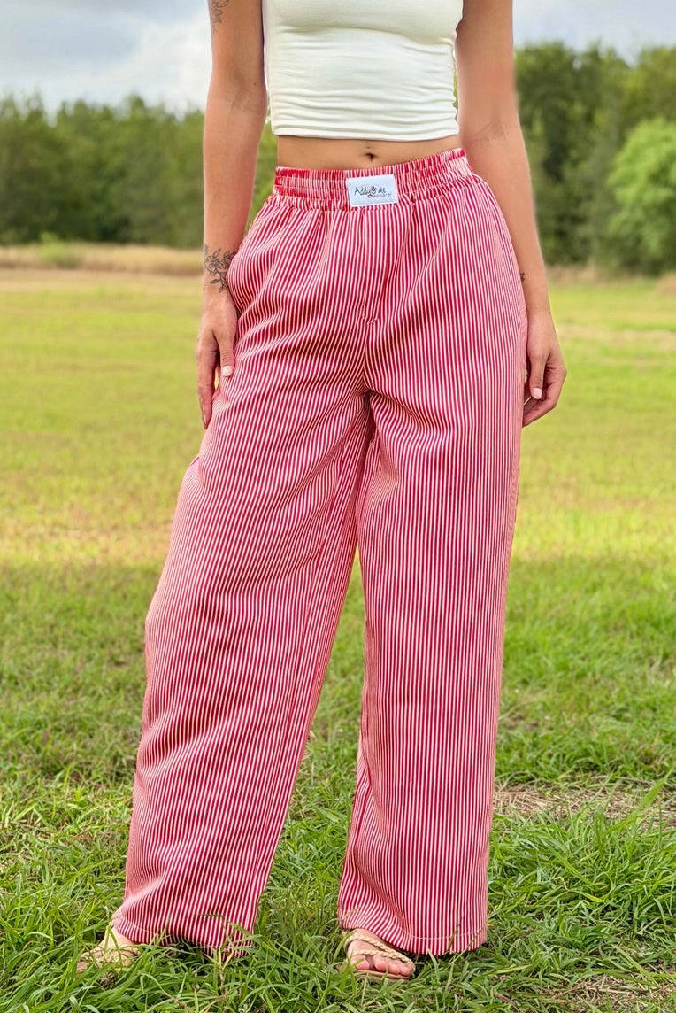 A person standing outdoors in a grassy field wearing loose, wide-leg pink pants with thin vertical darker pinstripes, a white cropped tank top, and sandals.