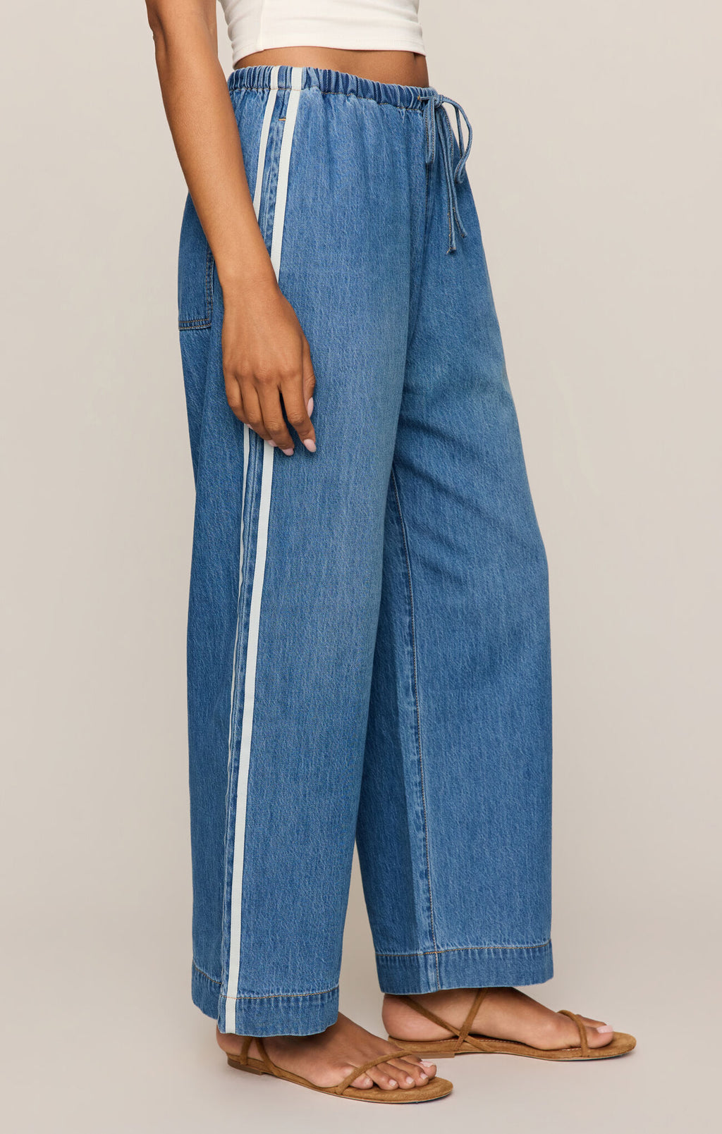 A person wearing wide-leg blue denim pants with a white side stripe and drawstring waist, paired with tan flat sandals against a plain light beige studio background.