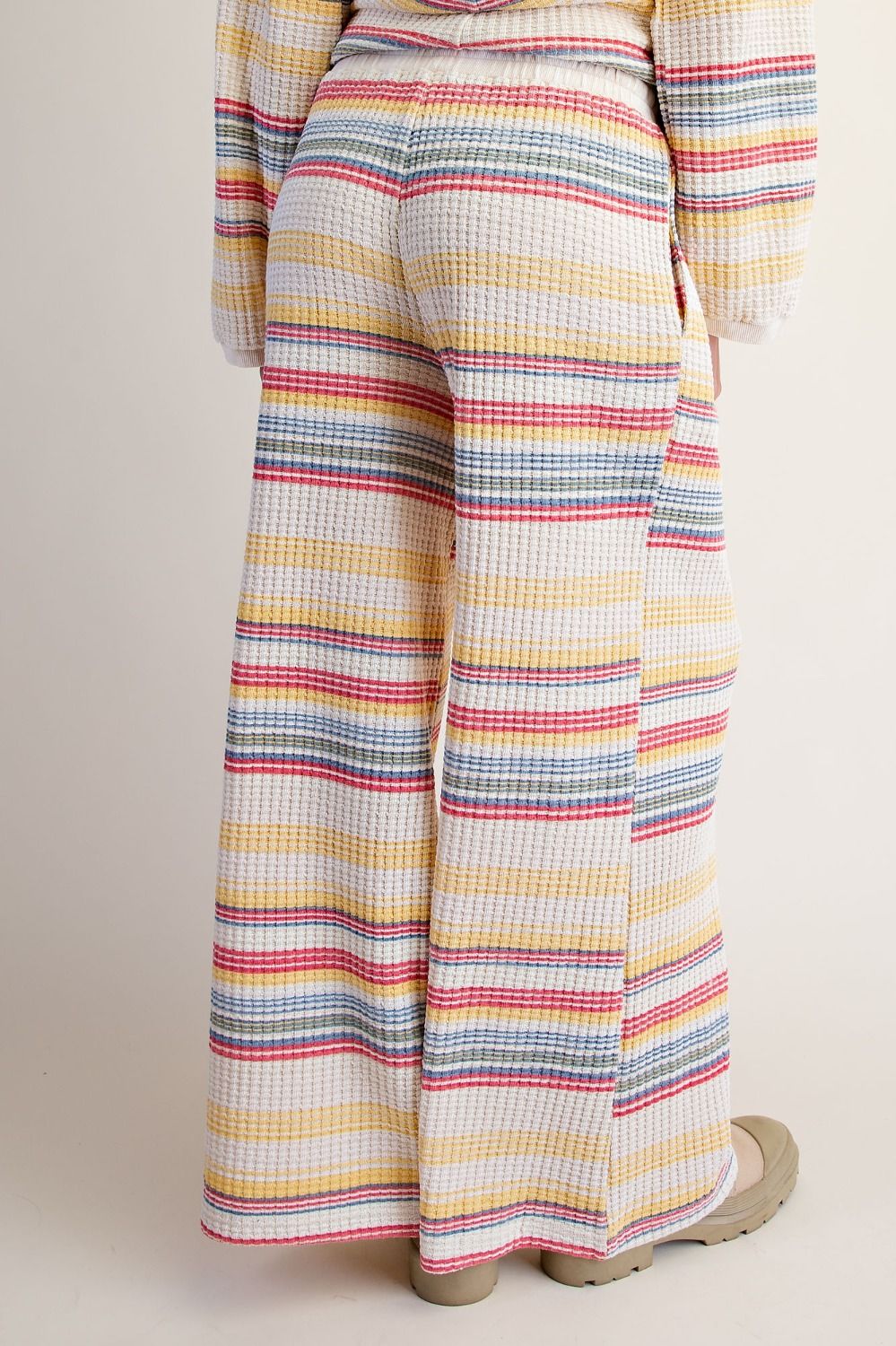 A person shown from the back wearing wide-leg waffle-knit pants with horizontal stripes in yellow, pink, blue, and gray on a cream base, paired with beige flat shoes against a plain light beige background.