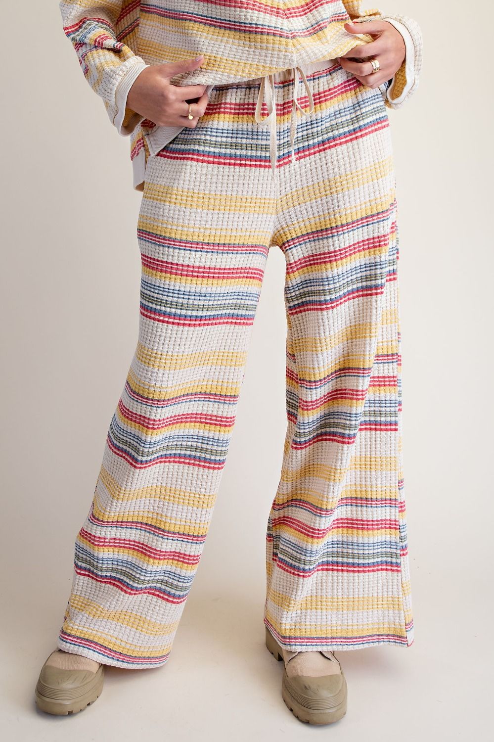 A person wearing wide-leg drawstring pants with horizontal multicolor stripes in yellow, red, blue, and gray on a cream base, paired with beige shoes against a plain beige studio background.